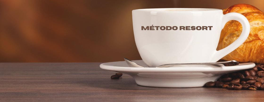 Xícara de café com a marca Método RESORT, ao lado de um croissant e grãos de café sobre a mesa